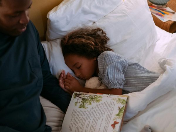 A father reading a bedtime story to his daughter, who is sleeping peacefully in bed.