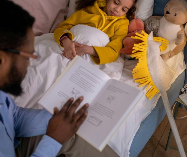 A father reading a bedtime story to his daughter tucked in bed with her stuffed toys.