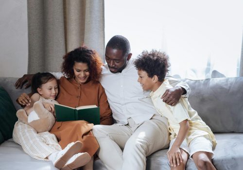 A loving family of four enjoys reading a book together in a cozy indoor setting.